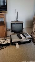 Panasonic 13-inch CRT TV with remote, Toshiba DVD player, Zenith VHS player, Zenith DTV tuner and all remotes together on carpeted floor.