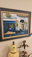 European street scene painting in ornate dark wooden frame with no glass, showing architectural details and shop window