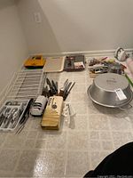 Overview of lot arrangement on floor showing cutlery trays, wood handled knives, wok pan, baking pan, kitchen gadgets including GE slicer and Salton warming tray.