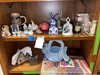 Shelf with assorted porcelain thimbles, ceramic windmills, Norman Rockwell bell without striker, candles, napkin holders, quarter maps, and various decorative items.