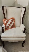 Full view of vintage wing chair with patchwork pillow on seat, showing wood frame and cream upholstery.
