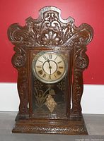 Front view of antique gingerbread clock showing carved wooden case, Roman numeral clock dial, and etched glass panel with visible decorative pendulum.