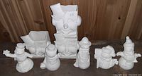 Photo of nine unpainted ceramic bisque snowmen pieces arranged on wooden surface, including seven small figures and two larger hollow head planters with hats.