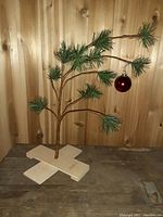 Photo showing small Charlie Brown Christmas tree with sparse pine branches and one red ornament hanging. Tree mounted on wooden cross base and set against wood paneling background.