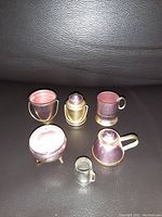 Photo showing six miniature metal vessels on a dark surface, including pitchers, cups and a container.
