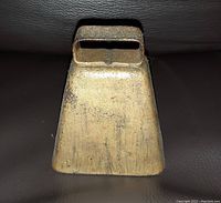 Front view of a rustic metal cowbell with natural patina placed on a dark brown surface.