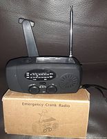 Photo shows a dark gray emergency crank radio with extended telescoping antenna, tuning and volume dials, speaker grill, and crank handle. The radio rests on the original cardboard box labeled 'Emergency Crank Radio'.