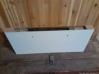White floating shelf lying against a wooden wall surface, showing the top and side edges of the shelf with mounting hardware partially visible in packaging.