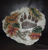 Stone decorative piece showing a bear paw print with colorful autumn leaves and branches around it on a textured stone base.