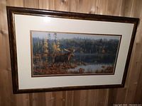 Framed James Hautman moose print hanging on wood panel wall, showing the entire artwork and frame.