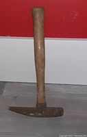 Pick hammer standing upright against a red wall and grey floor, showing the wooden handle and rusted metal hammer head.