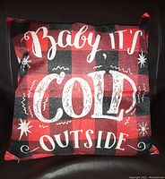 Front view of a square decorative pillow with red and black buffalo plaid design, white lettering saying 'Baby It's Cold Outside' with snowflake and star embellishments.