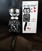 Black reproduction Kit-Cat clock next to its original box, showing cat figure with rolling eyes, bow tie, and wagging tail feature.