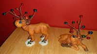 Photo of two wooden holiday moose decorations on a table against red background. One is standing and the other lying down, both with colored balls on antlers.