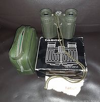 Green Tasco binoculars with attached gold chain next to green zippered case, cleaning cloth, manual, and original black Tasco box with white illustration, all placed on dark leather surface.