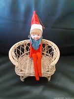 Wide shot of vintage Elf on the Shelf doll seated in vintage wicker doll chair on black background