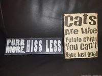 Photo of two wood block signs on a dark surface. One black sign reads 'Purr More, Hiss Less' in white bold letters. The other cream-colored sign reads 'Cats Are Like Potato Chips You Can't Have Just One' in distressed black text.