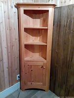 Front view of the wooden corner cabinet showing three open shelves and a closed door at the bottom.