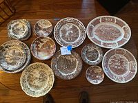 Photo showing all 14 assorted brown decorative plates in various sizes with scenes of historic buildings, villages, landscapes, and ships.