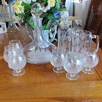 Full set of etched glassware including decanter, six stemmed glasses, and two mugs displayed on a wood table.