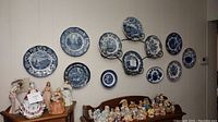 Wide view of 11 of the blue and white plates hung on the wall alongside figurines below, showing various scenic and floral designs in blue and white.