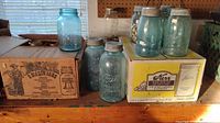 Wide view showing large blue Ball mason jar and boxes containing miscellaneous clear jars on a wooden surface