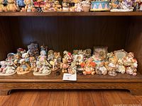Photo shows a shelf with multiple Cherished Teddies ceramic figurines arranged in a row, featuring bears in different settings and costumes, some holding props.