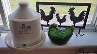 Photo showing white ceramic chicken waterer crock with chicken silhouette, green glass chicken basket, and metal rooster plaque with hooks in background