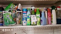 Shelf holding multiple assorted cleaning supplies including sprays, liquid cleaners, and a white basket with sponges