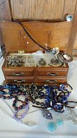 Wooden jewelry box open showing drawers and compartments with assorted costume jewelry inside and necklaces draped in front.
