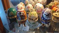 View of ten egg trinket boxes with floral and figurative designs, including a green one with a clock inside 