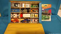 Top-down view of the opened collector's case displaying 12 vintage Hot Wheels and Matchbox cars. Also shows green Hot Wheels truck in original packaging beside the case.