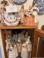 Photo of multiple porcelain ladies figurines displayed on top of and inside a wooden cabinet, showing different styles and poses in varied pastel colors.