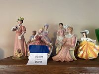 Wide shot showing all seven porcelain lady figurines in a row on a shelf against a beige wall.