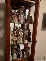 Photo showing three shelves of a wooden glass display case filled with various porcelain lady figurines in vintage attire, highlighting the varied details and accessories.