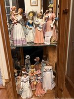Full view of two glass cabinet shelves filled with porcelain lady figurines dressed in historical and vintage style clothing, holding infants and accessories like hats, umbrellas, and paper.