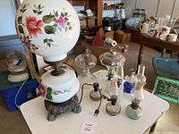 Photo showing entire lot of lamps on a white surface in a room with furniture in background. Large floral painted hurricane lamp with metal base and electric cord, plus seven smaller glass kerosene lamps of various styles.