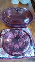 Top-down view of two covered casserole dishes on a wooden table, showing the larger Pyrex dish with lid and smaller Visions dish with lid placed on a quilted floral bag.