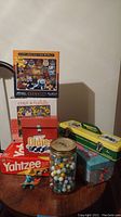 Full view of lot items arranged on table including two cat puzzles, Yahtzee game, Oldies CDs, vintage marbles jar, John Deere tin, Smurf figurine, Gameboy touchscreen visible in background