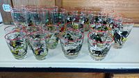 22 vintage rodeo and Old West glassware pieces arranged in rows, showing colorful scenes of bull riding, cattle, and covered wagons with red rimmed tops.