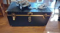 Black rectangular storage chest with brass hardware, riveted edges, and handles, with items on top including Dyson handheld vacuum body and power strip.