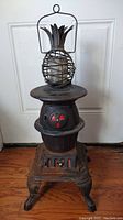Front view of the vintage cast iron stove with pineapple candle holder on top, showing the ornate legs and red vent accents.