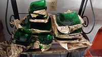 Overall view of stacked green glass plates, bowls, and vases partially wrapped in newspaper on metal shelf.
