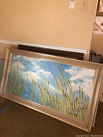 Wide view of the largest framed painting showing tall grasses with blue sky and white clouds, leaning against wall