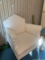 White upholstered armchair with cushioned seat and backrest, white removable arm covers, wooden legs with minor scratches.