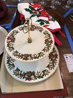 Two-tier ceramic serving tray decorated with holly leaves, red berries, gold bells, and ribbon garland, with a gold handle, set on top of a red and white Christmas dish towel with holiday designs.