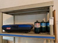 Photo of blue Coleman camping stove closed on shelf alongside four propane containers, one Coleman branded, three others smaller and green-blue colored.