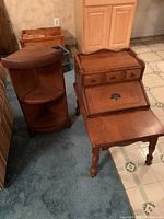 Photo of two vintage wooden tables: a corner stand with two shelves and a side table with a drop-front compartment and drawers, placed on carpet and linoleum flooring.