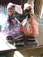 Two vintage-style music box dolls on rotating bases. One doll wears a purple floral dress and holds a lace parasol; the other wears a multicolor floral dress with ruffle accents. Five embroidered purses are placed in front of the dolls.
