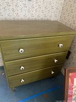 Front view of green dresser showing three drawers with white ceramic pulls, surface wear visible on top and drawers.